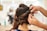A person is styling long brown hair, creating an intricate updo with curls being pinned into place. The focus is on the hands of the stylist as they carefully arrange the hair, with a blurred salon background.
