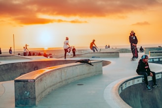 Skateboarders are enjoying an evening at a skate park, performing tricks and socializing under a vibrant sunset. The skate park features concrete ramps and bowls, and several people are seen engaging in dynamic activities.