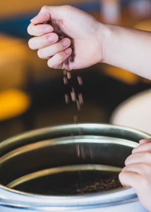 A hand holds coffee beans, allowing them to fall into a metal container below. The background is blurred, emphasizing the motion of the dropping beans.