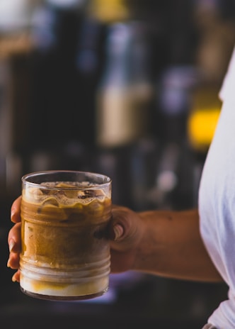 Close-up of a hand holding a glass of iced coffee with condensation droplets against a dark green backdrop.