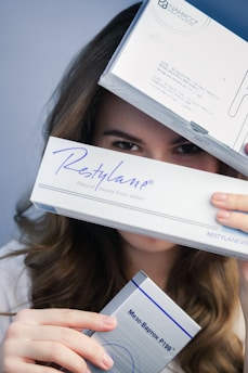 A person is holding three boxes, with focus on the box labeled 'Restylane'. The upper box partially covers their face, revealing only the eyes. The middle box prominently displays the 'Restylane' branding, and the bottom box has text in another language.