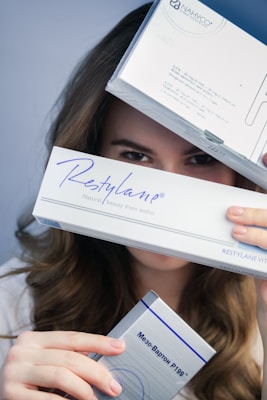 A person is holding three boxes, with focus on the box labeled 'Restylane'. The upper box partially covers their face, revealing only the eyes. The middle box prominently displays the 'Restylane' branding, and the bottom box has text in another language.