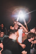 A group of people appears to be enjoying an energetic event or gathering. The main focus is on a person in a white shirt and cap, surrounded by others who are cheering, smiling, and showing excitement. Bright lights and possibly a lens flare are visible in the background, adding a dramatic effect to the scene.
