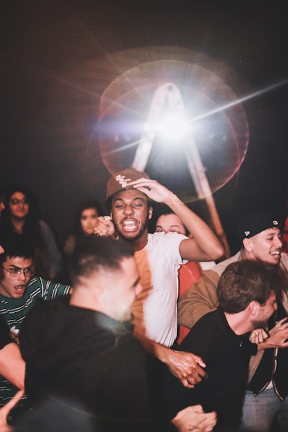 A group of people appears to be enjoying an energetic event or gathering. The main focus is on a person in a white shirt and cap, surrounded by others who are cheering, smiling, and showing excitement. Bright lights and possibly a lens flare are visible in the background, adding a dramatic effect to the scene.