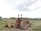 A red tractor with large wheels is parked on a dirt field. It is equipped with a plowing attachment at the back, which is partially loaded with soil. The landscape includes green foliage and trees in the background under an overcast sky.