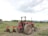 A red tractor with large wheels is parked on a dirt field. It is equipped with a plowing attachment at the back, which is partially loaded with soil. The landscape includes green foliage and trees in the background under an overcast sky.