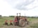 A red tractor with large wheels is parked on a dirt field. It is equipped with a plowing attachment at the back, which is partially loaded with soil. The landscape includes green foliage and trees in the background under an overcast sky.