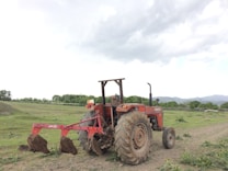 A red tractor with large wheels is parked on a dirt field. It is equipped with a plowing attachment at the back, which is partially loaded with soil. The landscape includes green foliage and trees in the background under an overcast sky.