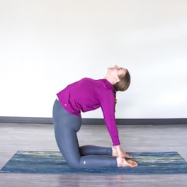 A person is performing a yoga pose on a yoga mat indoors. They are kneeling, leaning back with hands on their heels. The individual is wearing a purple long-sleeve top and gray leggings. The room has a light grey floor and an off-white wall.