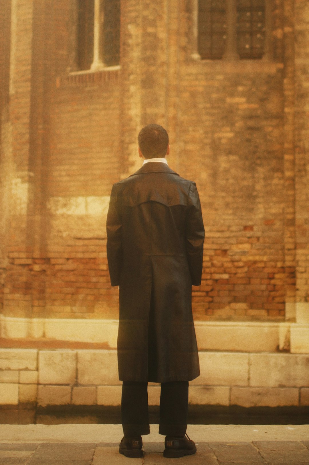 man in black suit standing near brown brick wall