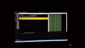 The image features a computer screen displaying a coding environment. The screen shows a text editor with various files and folders listed on the left-hand side, while the main coding area is open with a command line interface. There are green text outputs typical of command line operations performed on a black background.