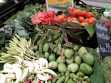 A vibrant display of fresh produce featuring red tomatoes, pink flowers, and green mangoes arranged on a market stall. The scene includes a mix of flowers, leafy greens, and vegetables, set against a backdrop of banana leaves. A chalkboard sign with writing in Thai is visible alongside the produce.