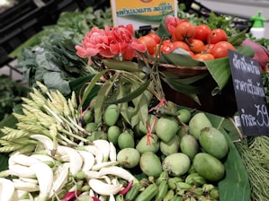 A vibrant display of fresh produce featuring red tomatoes, pink flowers, and green mangoes arranged on a market stall. The scene includes a mix of flowers, leafy greens, and vegetables, set against a backdrop of banana leaves. A chalkboard sign with writing in Thai is visible alongside the produce.