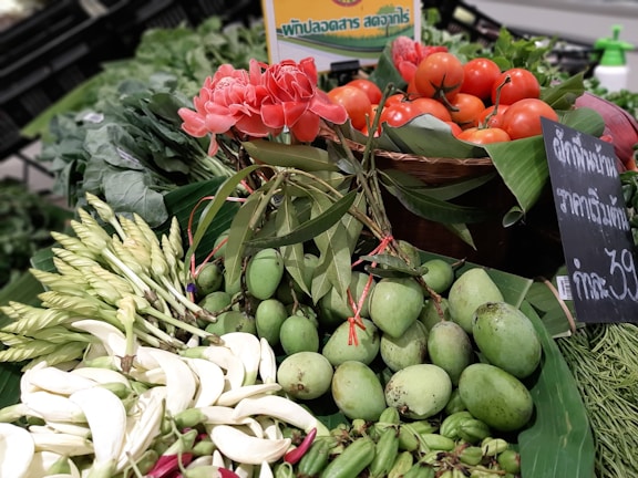 A vibrant display of fresh produce featuring red tomatoes, pink flowers, and green mangoes arranged on a market stall. The scene includes a mix of flowers, leafy greens, and vegetables, set against a backdrop of banana leaves. A chalkboard sign with writing in Thai is visible alongside the produce.