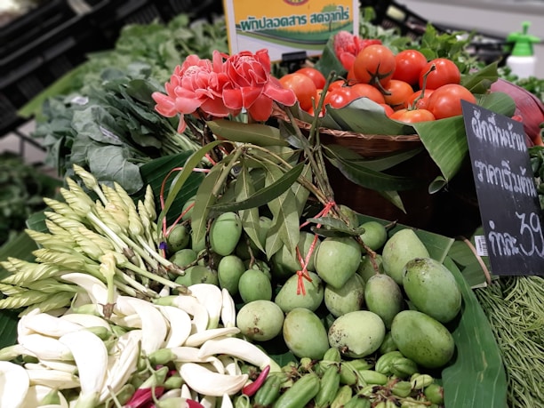A vibrant display of fresh produce featuring red tomatoes, pink flowers, and green mangoes arranged on a market stall. The scene includes a mix of flowers, leafy greens, and vegetables, set against a backdrop of banana leaves. A chalkboard sign with writing in Thai is visible alongside the produce.