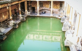 A historic bathhouse featuring a large rectangular pool filled with green water, surrounded by stone columns and walls. The architecture reflects ancient design with arches and well-preserved stone structures. Natural light casts reflections on the water, creating a serene atmosphere.