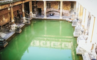 A historic bathhouse featuring a large rectangular pool filled with green water, surrounded by stone columns and walls. The architecture reflects ancient design with arches and well-preserved stone structures. Natural light casts reflections on the water, creating a serene atmosphere.