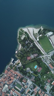 An aerial view of a coastal area with a blend of urban and natural elements. The top left features a large body of water with a shoreline curving inward, surrounded by a grassy park area with pathways. To the right, there are sports fields, including a soccer field and a tennis court, as well as a building complex. The bottom section of the image shows a densely built residential area with multiple red-roofed structures.
