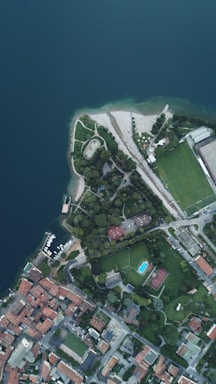 An aerial view of a coastal area with a blend of urban and natural elements. The top left features a large body of water with a shoreline curving inward, surrounded by a grassy park area with pathways. To the right, there are sports fields, including a soccer field and a tennis court, as well as a building complex. The bottom section of the image shows a densely built residential area with multiple red-roofed structures.