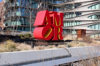 A large red sculpture spelling 'AMOR', placed in an outdoor urban setting with modern architecture in the background. The area around the sculpture is landscaped with grass and bare trees.