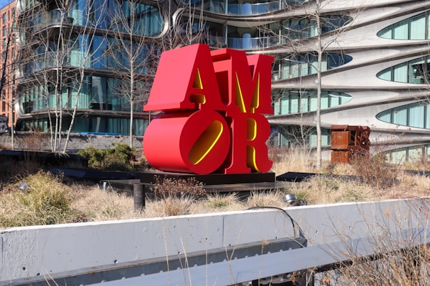 A large red sculpture spelling 'AMOR', placed in an outdoor urban setting with modern architecture in the background. The area around the sculpture is landscaped with grass and bare trees.