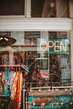 A shop window display with an eclectic collection of items, including colorful scarves, a painting, and a dark jacket adorned with a wolf-like face. The word 'OPEN' is illuminated in neon, indicating that the shop is welcoming customers. Reflections on the glass add a slightly urban feel.