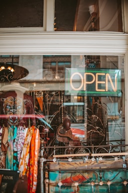 A shop window display with an eclectic collection of items, including colorful scarves, a painting, and a dark jacket adorned with a wolf-like face. The word 'OPEN' is illuminated in neon, indicating that the shop is welcoming customers. Reflections on the glass add a slightly urban feel.