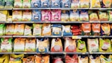 A grocery store aisle displays a variety of rice bags stacked neatly on shelves. These bags are of different brands and come in various colors and sizes. The packaging features colorful designs with images of rice and labels in multiple languages, indicating the type of rice and branding.
