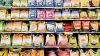 A grocery store aisle displays a variety of rice bags stacked neatly on shelves. These bags are of different brands and come in various colors and sizes. The packaging features colorful designs with images of rice and labels in multiple languages, indicating the type of rice and branding.