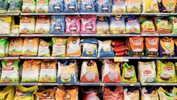 A grocery store aisle displays a variety of rice bags stacked neatly on shelves. These bags are of different brands and come in various colors and sizes. The packaging features colorful designs with images of rice and labels in multiple languages, indicating the type of rice and branding.