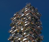 A tall, modern building with a unique design featuring numerous balconies covered with various types of plants and trees. The structure incorporates a vertical forest concept, blending urban architecture with greenery. The building's exterior is characterized by a mix of earthy tones from the plants and sleek gray materials.