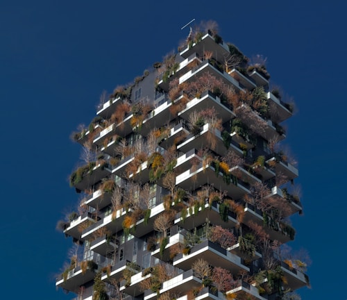 A tall, modern building with a unique design featuring numerous balconies covered with various types of plants and trees. The structure incorporates a vertical forest concept, blending urban architecture with greenery. The building's exterior is characterized by a mix of earthy tones from the plants and sleek gray materials.
