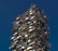 A tall, modern building with a unique design featuring numerous balconies covered with various types of plants and trees. The structure incorporates a vertical forest concept, blending urban architecture with greenery. The building's exterior is characterized by a mix of earthy tones from the plants and sleek gray materials.
