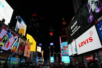 A bustling cityscape illuminated by numerous large, colorful digital billboards advertising brands like Levi's and Apple Music. The setting appears to be an urban environment, possibly a famous city square, with a crowd of people visible below the signs. Bright lights and advertisements create a lively atmosphere.