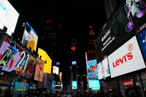 A bustling cityscape illuminated by numerous large, colorful digital billboards advertising brands like Levi's and Apple Music. The setting appears to be an urban environment, possibly a famous city square, with a crowd of people visible below the signs. Bright lights and advertisements create a lively atmosphere.