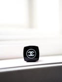 A close-up of a black makeup container with a recognizable logo on top. The container is placed on a light-colored surface, with bright light in the background creating a minimalist and elegant composition.
