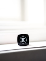 A close-up of a black makeup container with a recognizable logo on top. The container is placed on a light-colored surface, with bright light in the background creating a minimalist and elegant composition.