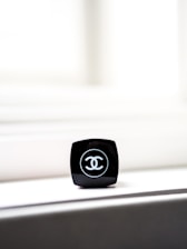 A close-up of a black makeup container with a recognizable logo on top. The container is placed on a light-colored surface, with bright light in the background creating a minimalist and elegant composition.