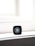 A close-up of a black makeup container with a recognizable logo on top. The container is placed on a light-colored surface, with bright light in the background creating a minimalist and elegant composition.