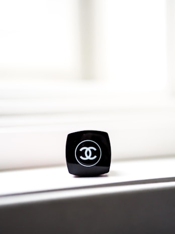 A close-up of a black makeup container with a recognizable logo on top. The container is placed on a light-colored surface, with bright light in the background creating a minimalist and elegant composition.