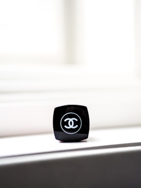 A close-up of a black makeup container with a recognizable logo on top. The container is placed on a light-colored surface, with bright light in the background creating a minimalist and elegant composition.