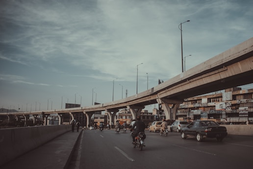 A multi-lane roadway with several vehicles, including cars and motorcycles, traveling in both directions. An elevated freeway or overpass stretches above the road, supported by large concrete pillars. The scene includes roadside buildings with numerous signboards.