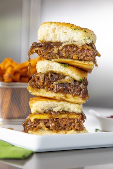 A stack of three mouth-watering sliders featuring caramelized onions and beef patties, placed on a white rectangular plate. A wire basket holding crispy sweet potato fries is visible in the background, adding depth and complementing the dish.