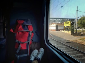 A view from inside a train compartment showing a red and black backpack next to a pair of white shoes with feet inside, resting on a seat. The train is at a station, with a sign visible outside the window indicating 'Thrissur' in multiple scripts. The background shows a railway platform, some greenery, and overhead electric lines under a clear sky.