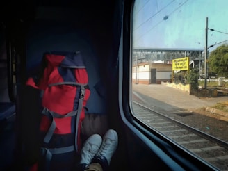 A view from inside a train compartment showing a red and black backpack next to a pair of white shoes with feet inside, resting on a seat. The train is at a station, with a sign visible outside the window indicating 'Thrissur' in multiple scripts. The background shows a railway platform, some greenery, and overhead electric lines under a clear sky.