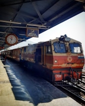 A train is stationed at a railway platform, with a clock displaying the time and a sign indicating a waiting room. The train is painted in red and cream colors, and there are a few people visible on the platform, which is covered with a metal roof.