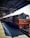 A train is stationed at a railway platform, with a clock displaying the time and a sign indicating a waiting room. The train is painted in red and cream colors, and there are a few people visible on the platform, which is covered with a metal roof.