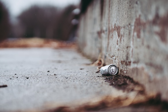 An empty aluminum can lies on the ground next to a rough concrete wall. The background appears blurry, suggesting a shallow depth of field. The setting looks like an outdoor urban environment, possibly a sidewalk or pathway.