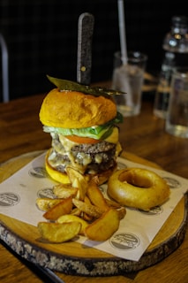 A gourmet burger with a knife stabbed through the top bun, layered with cheese, lettuce, and a beef patty, accompanied by thick-cut potato wedges and an onion ring on a wooden serving board. The scene is set against a rustic table, with glass bottles and a drinking glass in the background.