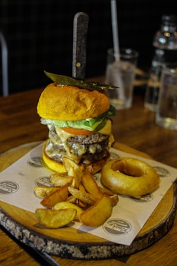 A gourmet burger with a knife stabbed through the top bun, layered with cheese, lettuce, and a beef patty, accompanied by thick-cut potato wedges and an onion ring on a wooden serving board. The scene is set against a rustic table, with glass bottles and a drinking glass in the background.